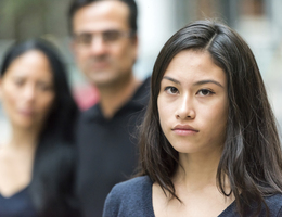 An unsmiling teenaged girl with her parents standing behind her.