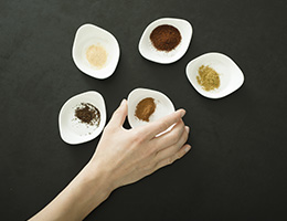 5 small white bowls of ground spices sit on a gray surface. A hand selects one of them. 