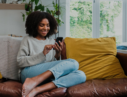 A young woman on a couch smiles as she looks at her phone screen.