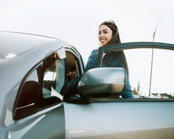 A woman stands near the open driver’s-side door of a compact car.