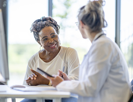 A woman sits beside a desk, talking with a seated doctor.