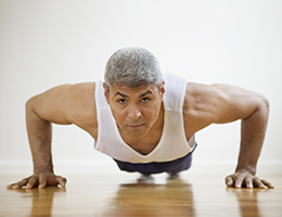 A man with graying hair wearing a white tank top stops at the lowest point of a pushup and looks up into the camera.